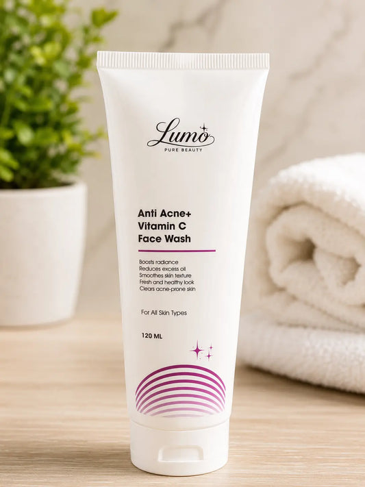 A white squeeze tube of Lumi Pure Beauty Anti Acne+ Vitamin C Face Wash, standing upright on a light wooden surface with a blurred green plant on the left and a folded white towel on the right.