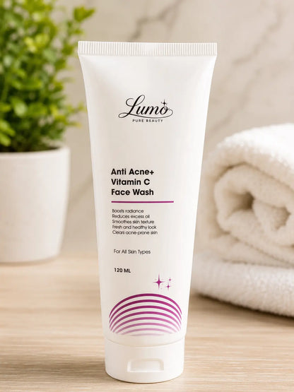 A white squeeze tube of Lumi Pure Beauty Anti Acne+ Vitamin C Face Wash, standing upright on a light wooden surface with a blurred green plant on the left and a folded white towel on the right.
