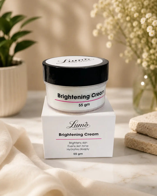 A jar of Lumi’s Brightening Cream sits on top of its white box, styled on a marble surface with soft beige fabric, a small potted plant, and dried flowers in the background.
