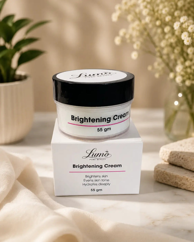 A jar of Lumi’s Brightening Cream sits on top of its white box, styled on a marble surface with soft beige fabric, a small potted plant, and dried flowers in the background.