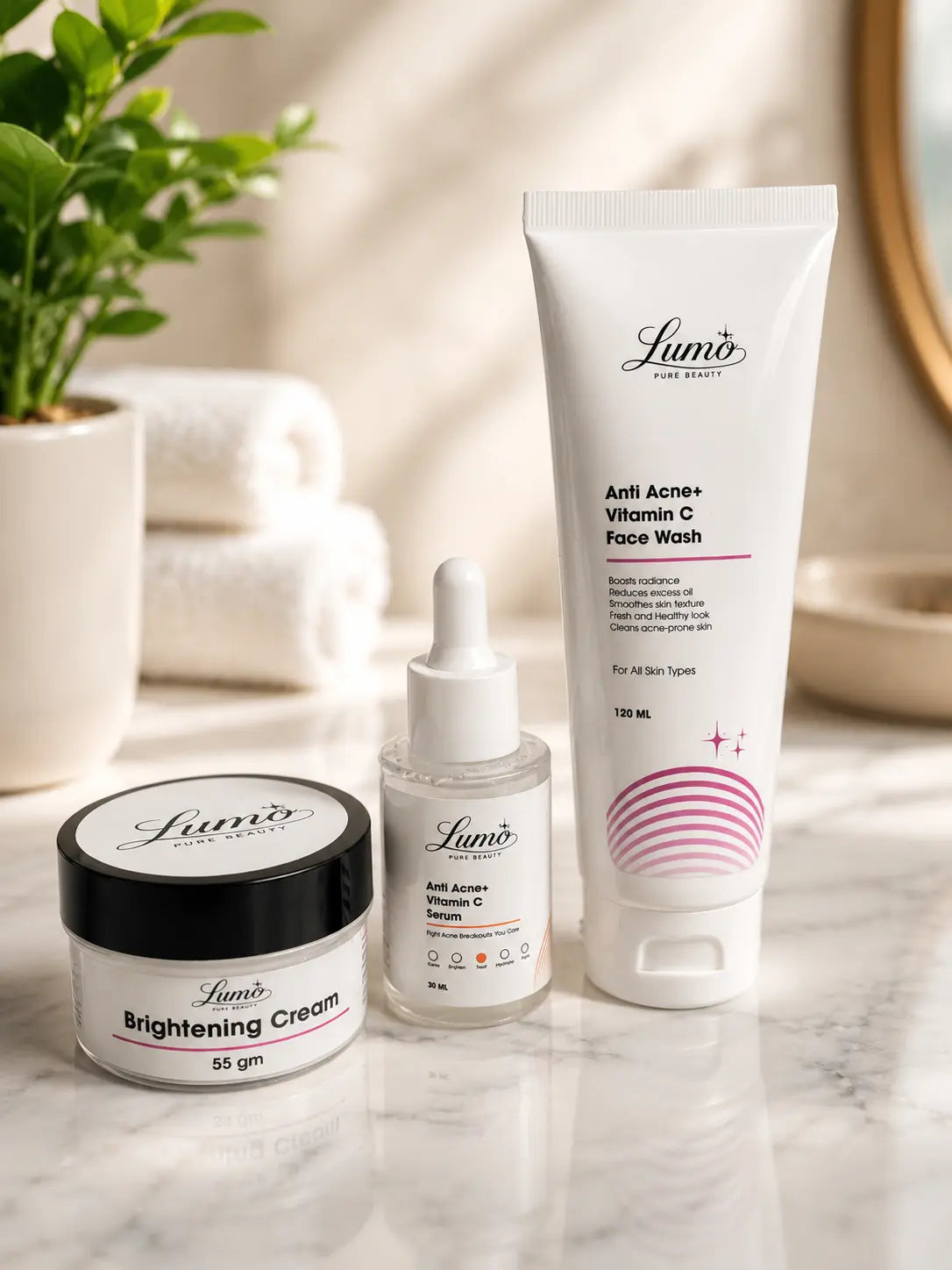 Three Lumo skincare products arranged on a marble surface: a brightening cream jar with a black lid, a small serum dropper bottle, and a larger vitamin C face wash tube, with folded towels and a plant in the softly lit background.