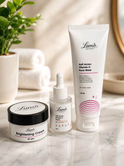 Three Lumo skincare products arranged on a marble surface: a brightening cream jar with a black lid, a small serum dropper bottle, and a larger vitamin C face wash tube, with folded towels and a plant in the softly lit background.