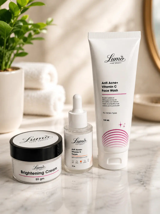 Three Lumo skincare products arranged on a marble surface: a brightening cream jar with a black lid, a small serum dropper bottle, and a larger vitamin C face wash tube, with folded towels and a plant in the softly lit background.